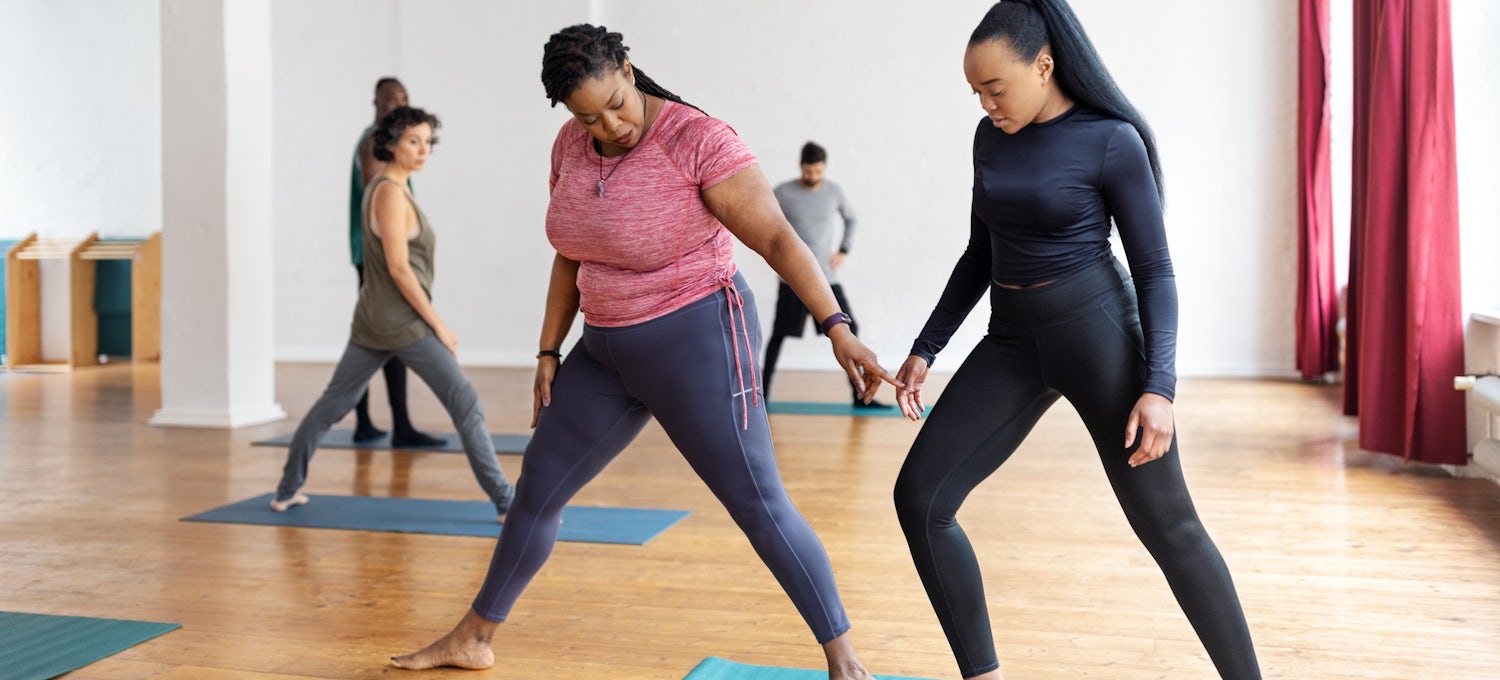 [Featured image] A fitness instructor assists a client in a group yoga class at a fitness studio.