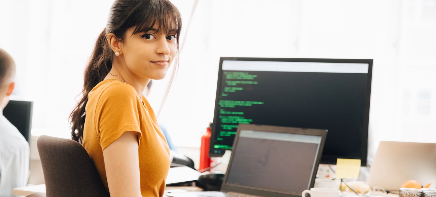 [Featured Image] A dark-haired programmer turns away from their laptop and computer monitor while learning how to code. 