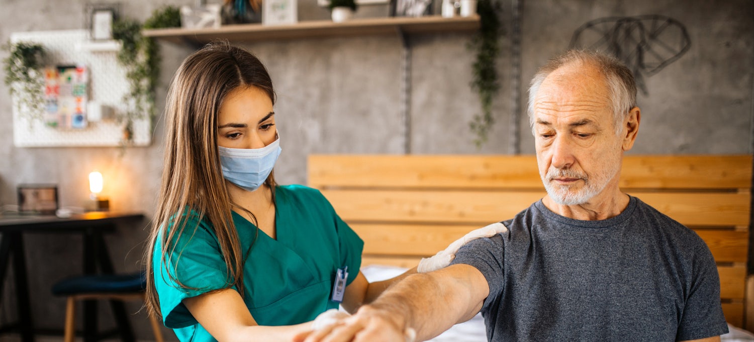 [Featured Image]:  A female home health aide, with long brown hair,  wearing a green uniform and a blue face covering, is taking care of a male patient in his home.  