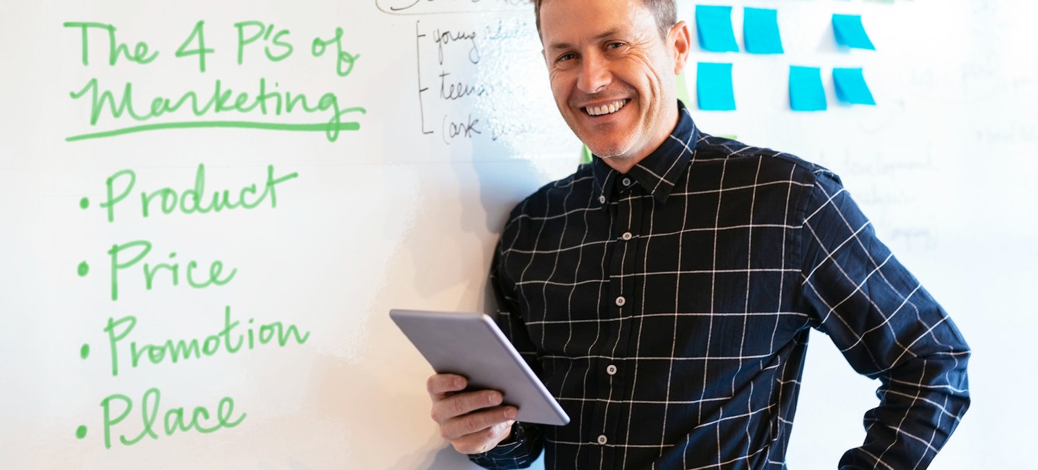 [Featured image] A man holding a tablet stands in front of a whiteboard where the 4 Ps of marketing are listed in green marker.