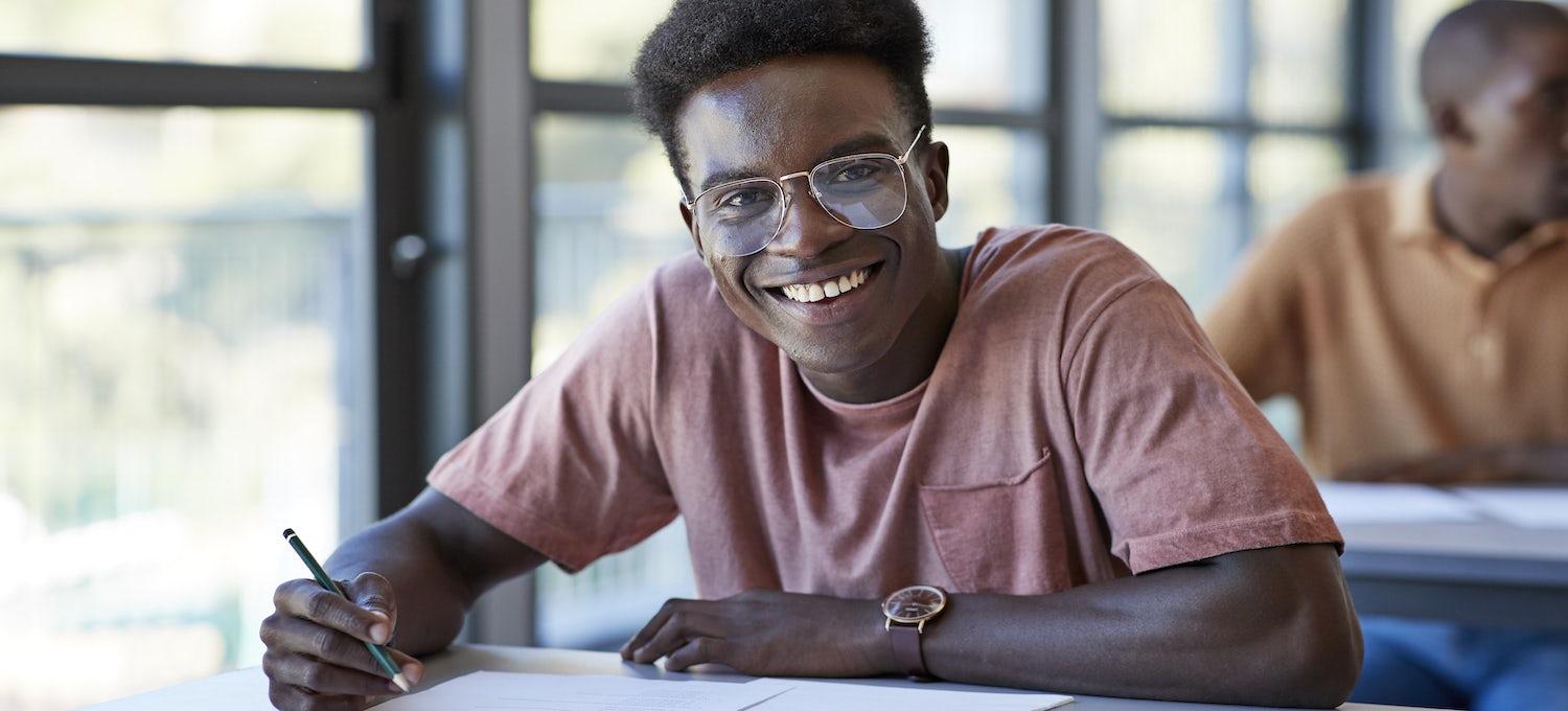 [Featured image] A young man wearing a rusty red t-shirt smiles at the camera as he takes a paper test with a pencil. 