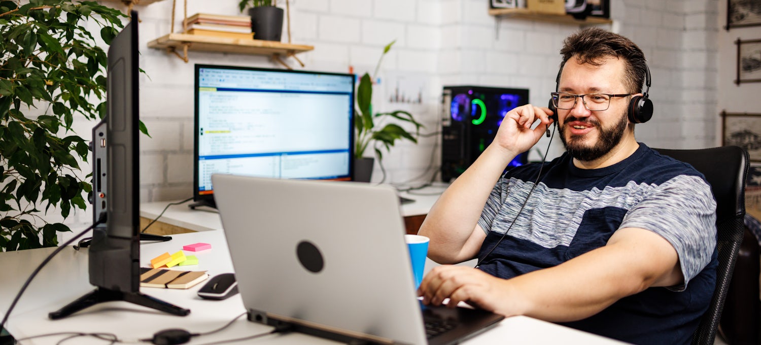[Featured image] A front-end web developer sits at a computer wearing a headset and chats with a client about creating a new website.