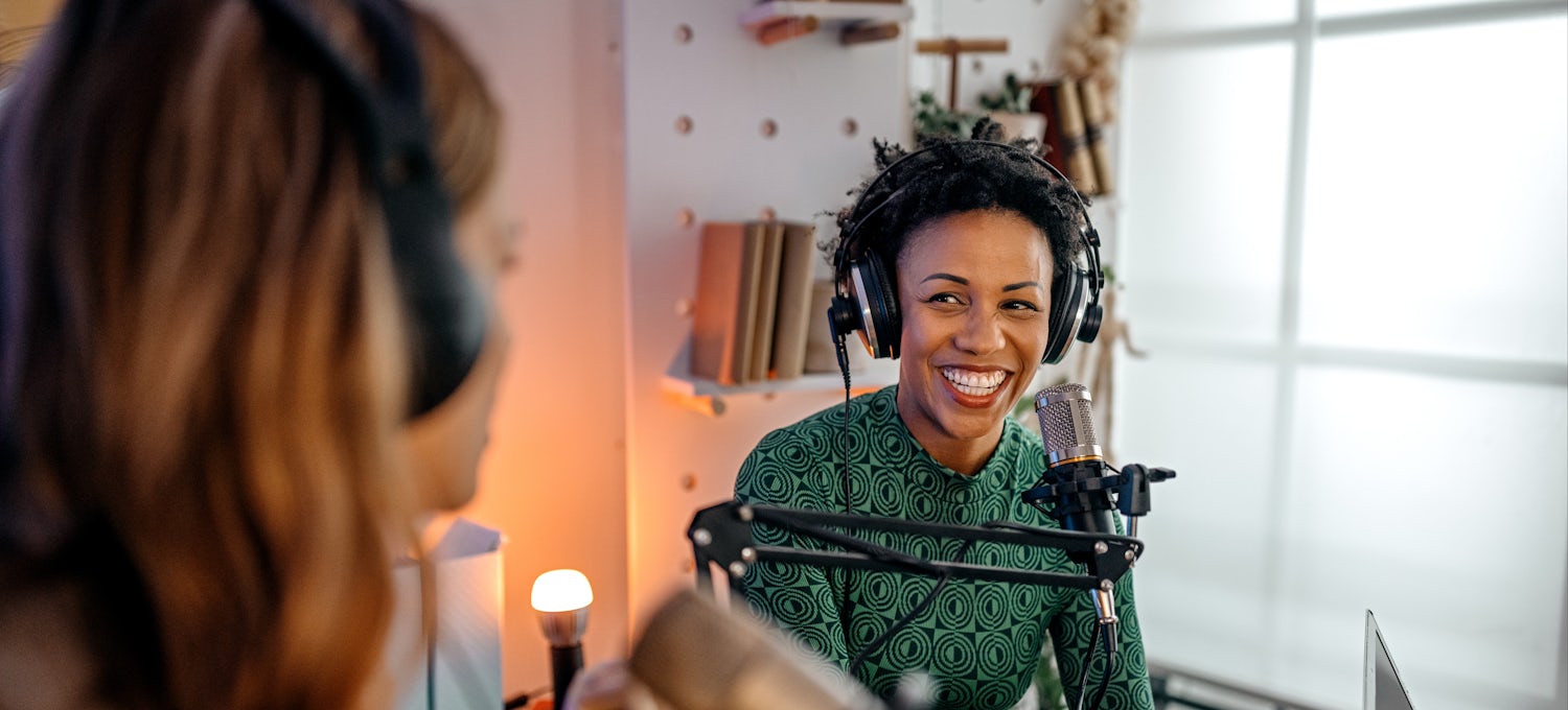 [Featured image] Two women wear over-ear headphones and record a podcast on microphones.
