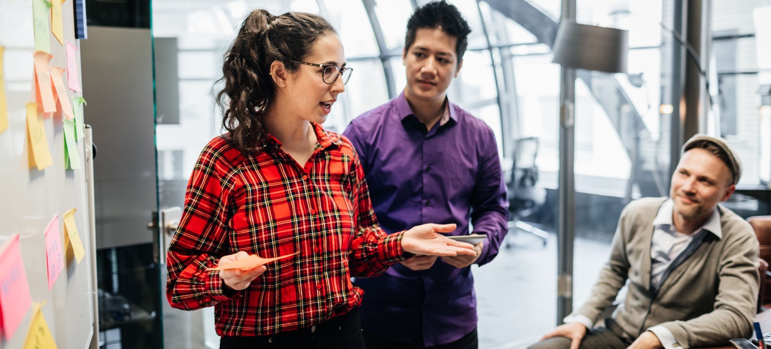 [Featured image] A Scrum Master in a red plaid shirt leads a meeting in front of a whiteboard covered in colorful sticky notes.