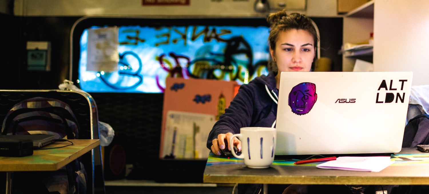 A young woman sits at a table in a cafe writing her resume on a laptop computer with stickers on it. She has a cup of coffee in front of her on the table.