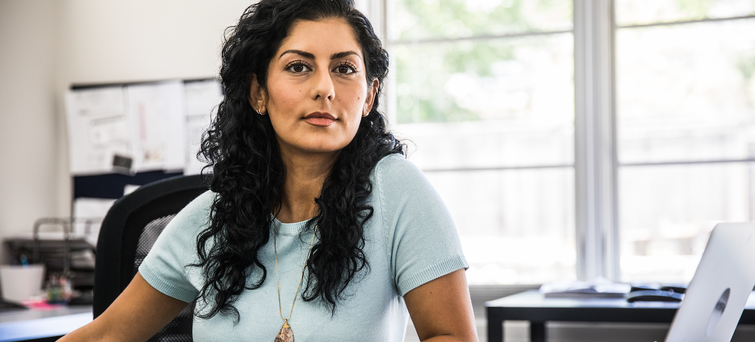 [Hauptbild] Eine Datenanalystin sitzt an ihrem Schreibtisch vor einem Laptop und schaut in die Kamera.
