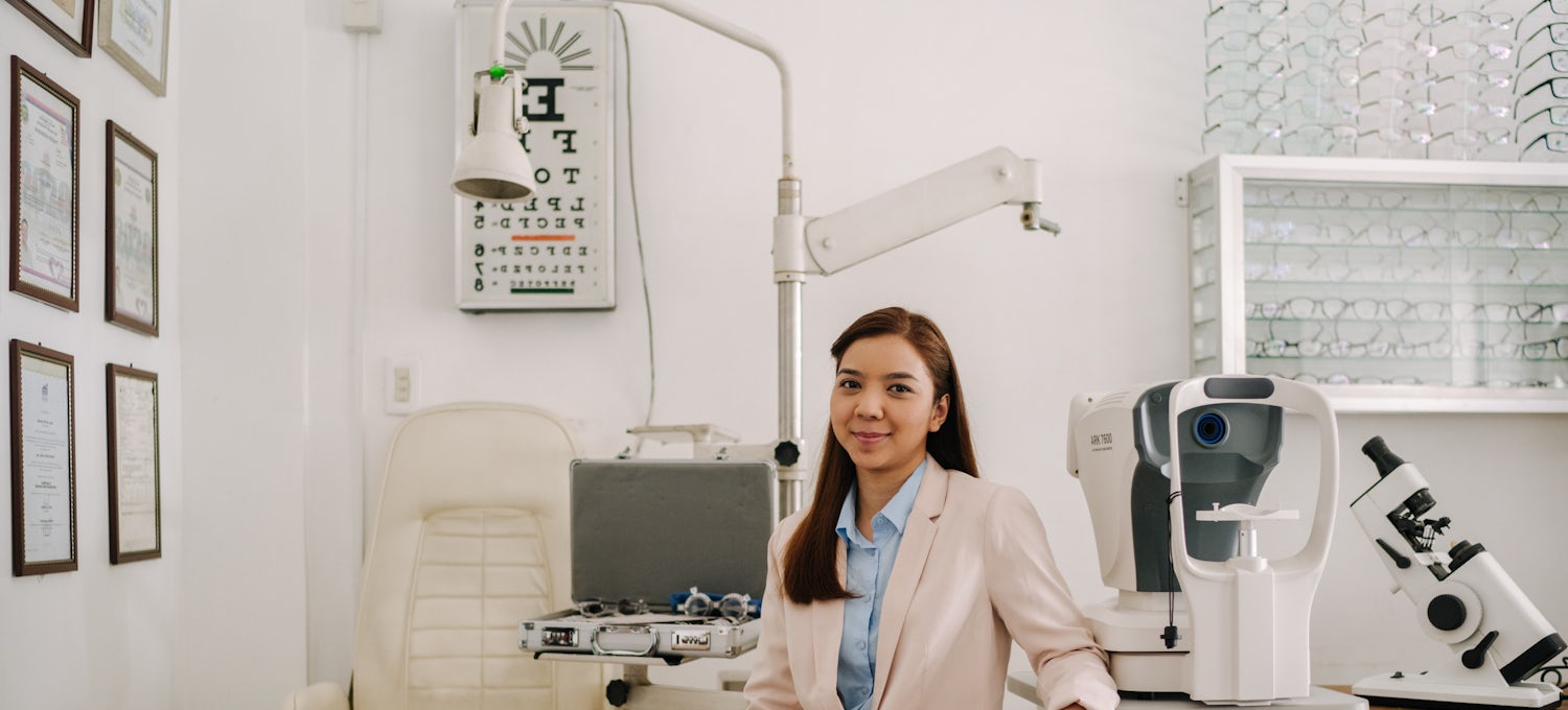 [Featured Image]:  Optometrist, wearing a white jacket and brown pants, sitting in the exam room, using health informatics skills.