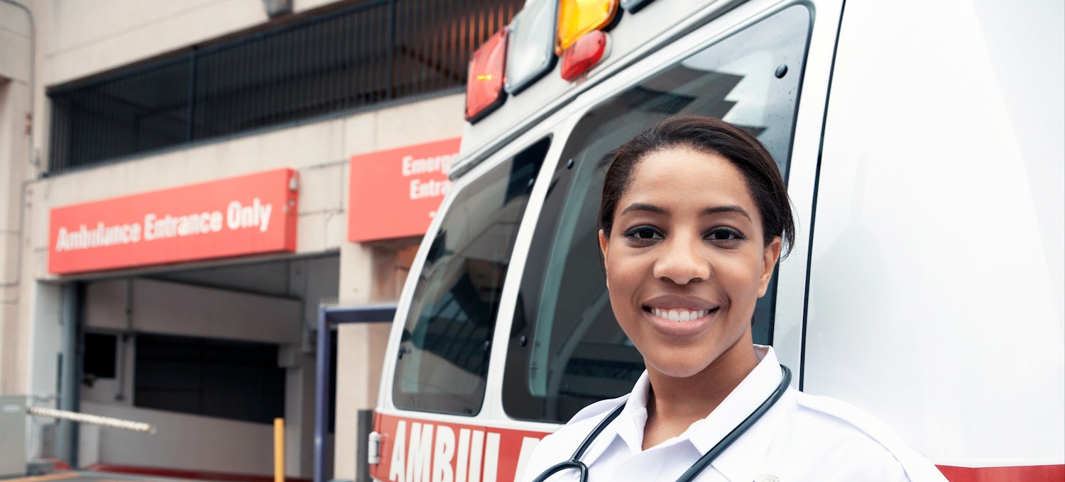 [Featured Image]: A woman with short dark hair, is wearing a white paramedic uniform. She has a stethoscope around her neck. She is standing in front of an ambulance at the entrance of the emergency room of a hospital. 