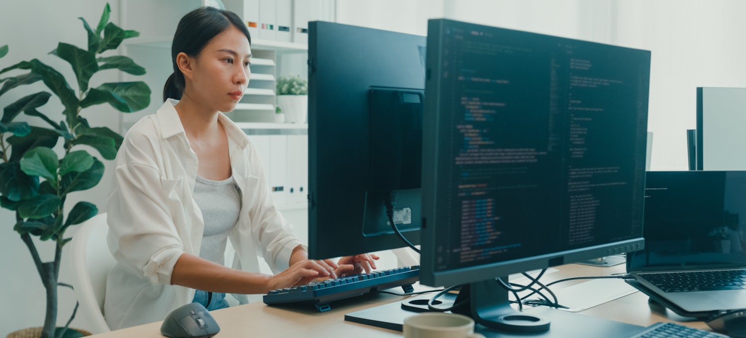A woman data scientist using a computer to write code sitting at a desk with multiple screens working at the office. 