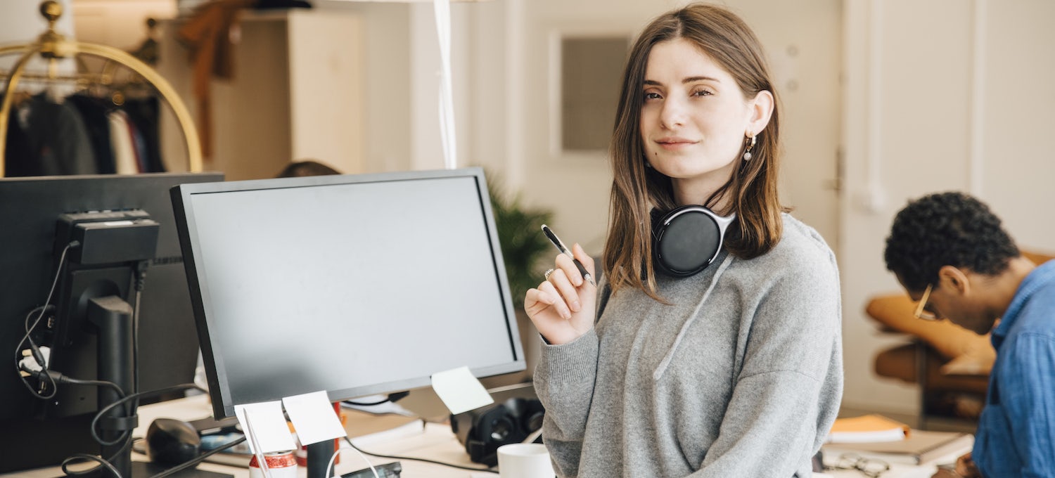 [Featured image] A data science degree student with over-the-ear headphones around her neck stands in front of a desk. 