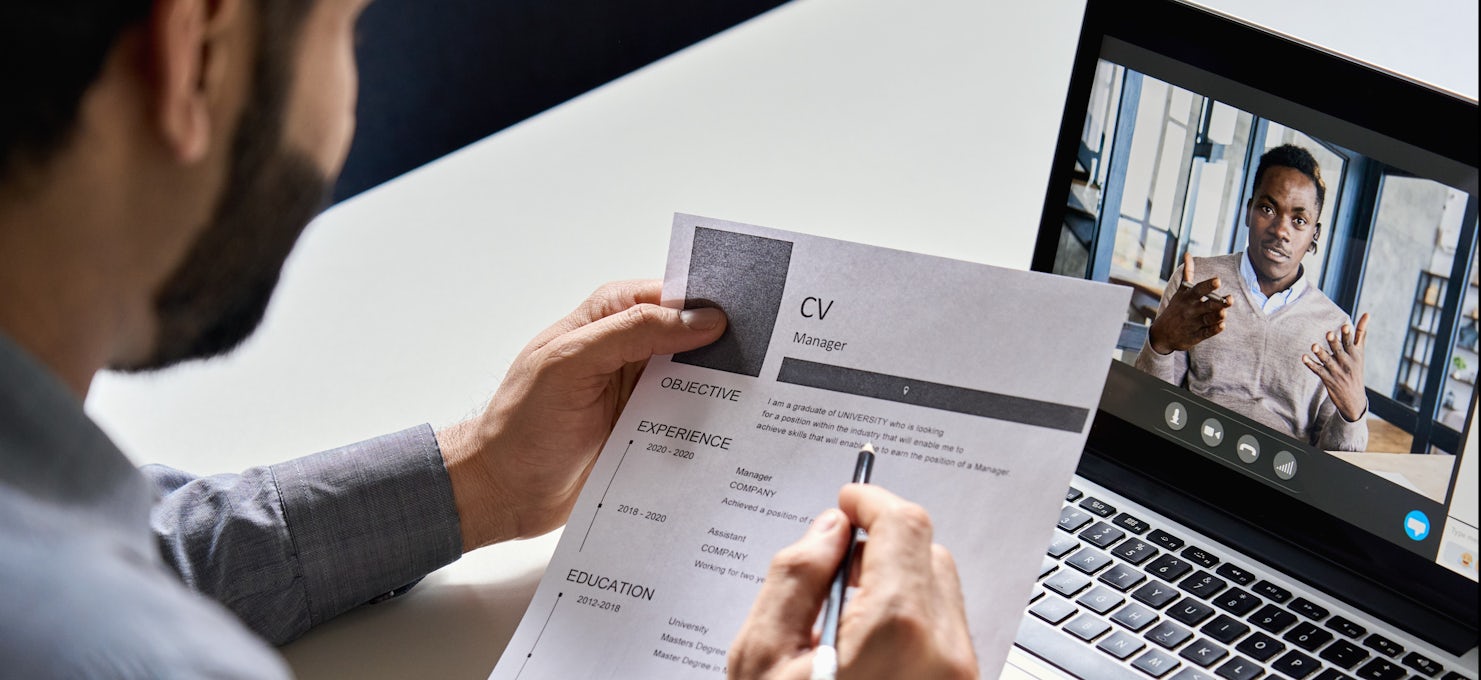[Featured image] A recruiter holds a printed CV and interviews a candidate virtually via laptop for a role in human resource management.