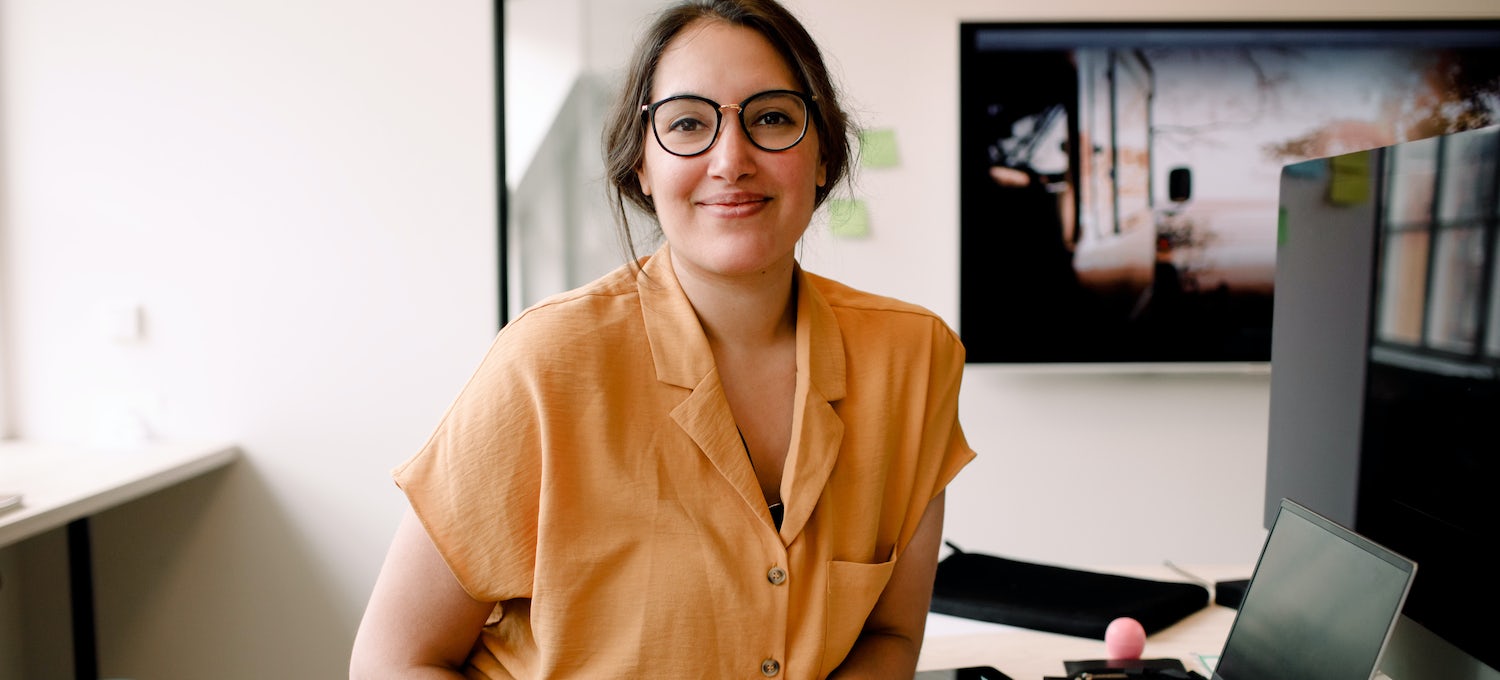 一位身穿黄色衬衫和眼镜的 UX 设计师坐在她的电脑前，对着镜头微笑