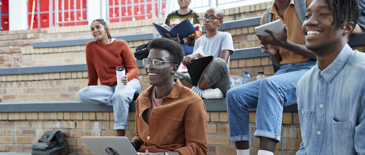 [Featured image] A group of six students sitting outside on a tiered brick stadium seating holding coffees, notebooks, and laptops, all smiling and looking toward a professor off-camera.