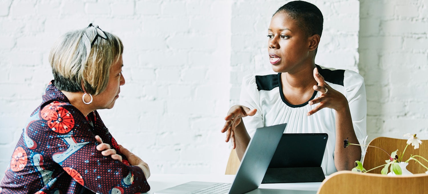 [Featured image] One woman speaks while the other listens, with a laptop in the foreground.
