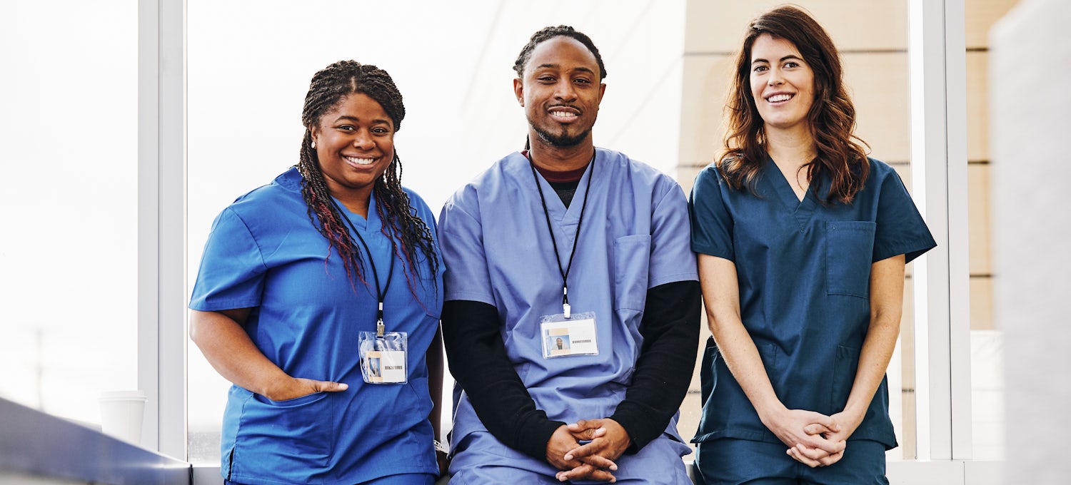 Three nurses, two female and one male all wearing blue scrubs, smile in a hospital. 