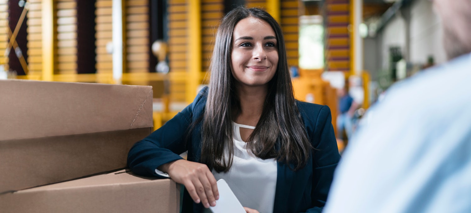 [Featured Image] A project manager in business casual clothing leans against cardboard boxes and talks to their coworker.