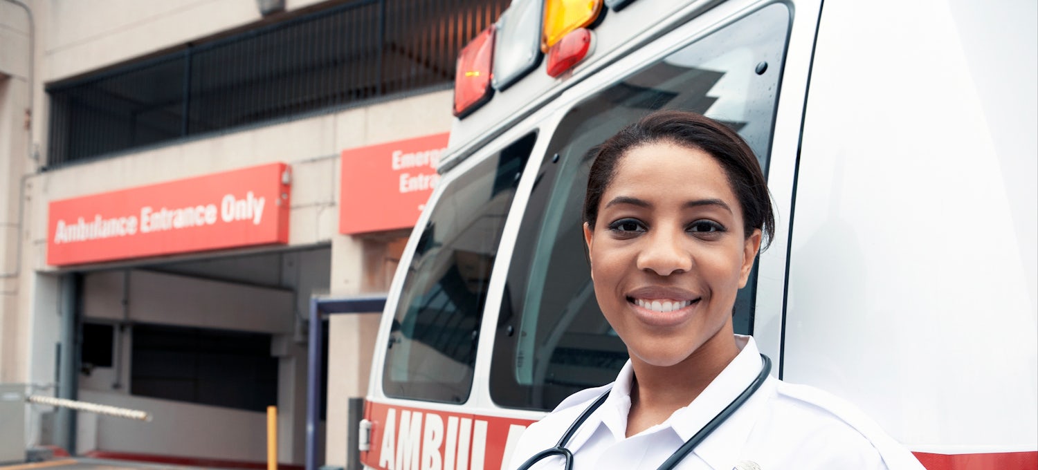 [Featured Image]:  A female paramedic, wearing a white uniform and a stethoscope around her neck, is standing in next to an ambulance, in front of an emergency room entrance. 