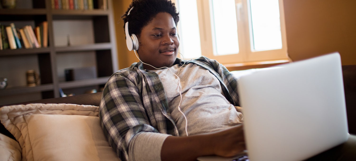 [Featured image] A young man in a plaid shirt wears headphones and works on his Associate of Science degree on his laptop.