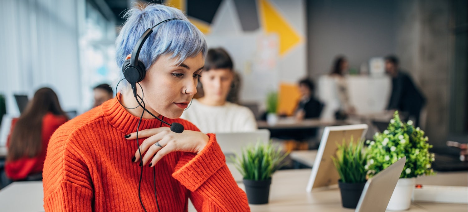 [Featured image] A person in an orange sweater and a blue pixie cut brainstorms character design ideas on their laptop in a co-working space.
