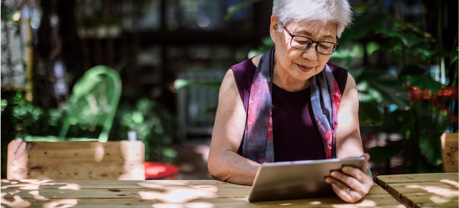 An older woman sits outside at a wooden table reading a book on her tablet device.