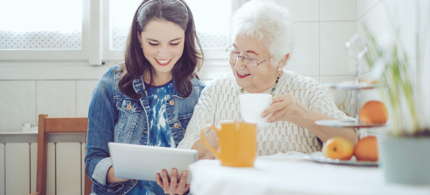 [Featured Image] A young woman sits in a kitchen with a tablet and volunteers with a senior citizen to help improve her service orientation skills.
