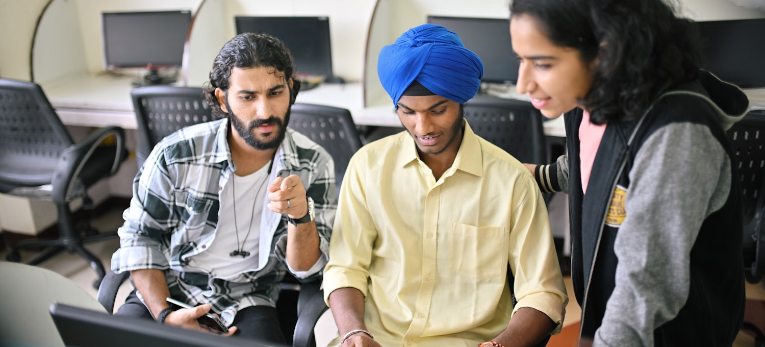 [Featured Image] Two seated students and one standing student look at a computer monitor and discuss the meaning and features of system programming.