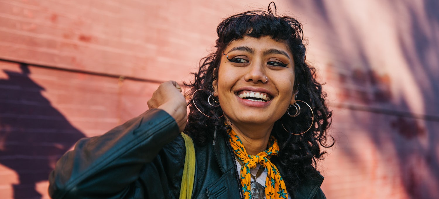 [Featured image] A young woman with curly black hair and big hoop earrings, wearing a black jacket and a color scarf, smiles at someone off-camera. 