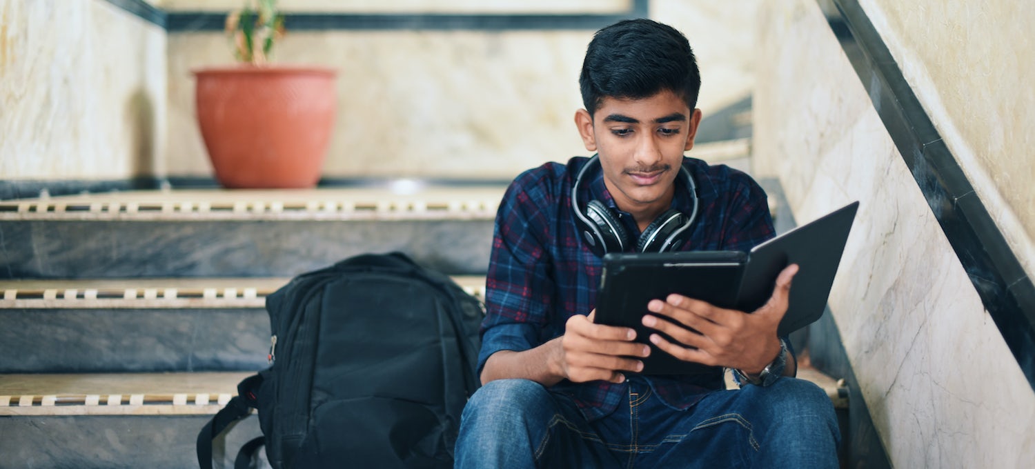 [Imagen destacada] Un estudiante universitario sentado en unas escaleras mirando su tableta. Su mochila está a su lado.