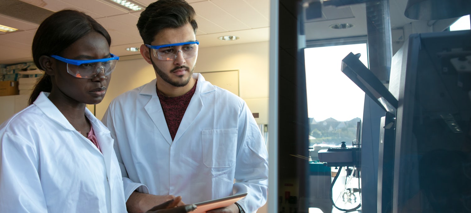 [Featured Image]: A female, with black hair, wearing a white uniform and blue goggles, and a male with black hair, wearing a white uniform and blue goggles. They are working in a lab, holding, writing and analyzing an experiment. 