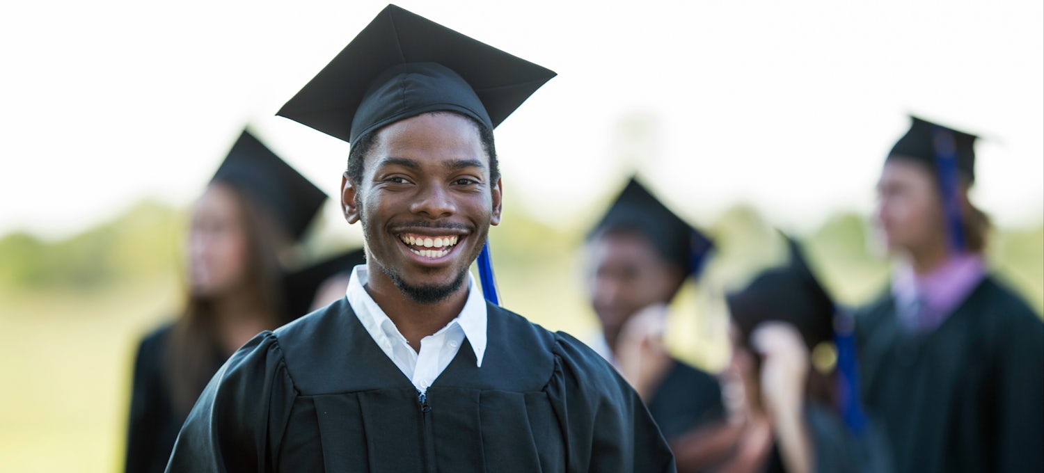 [Featured Image]:  A man wearing a black cap and gown smiles as he graduates as a social science major.