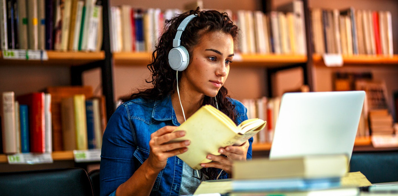 [Featured Image] A college student wears headphones and sits at a library table with a book in her hands and a laptop in front of her.