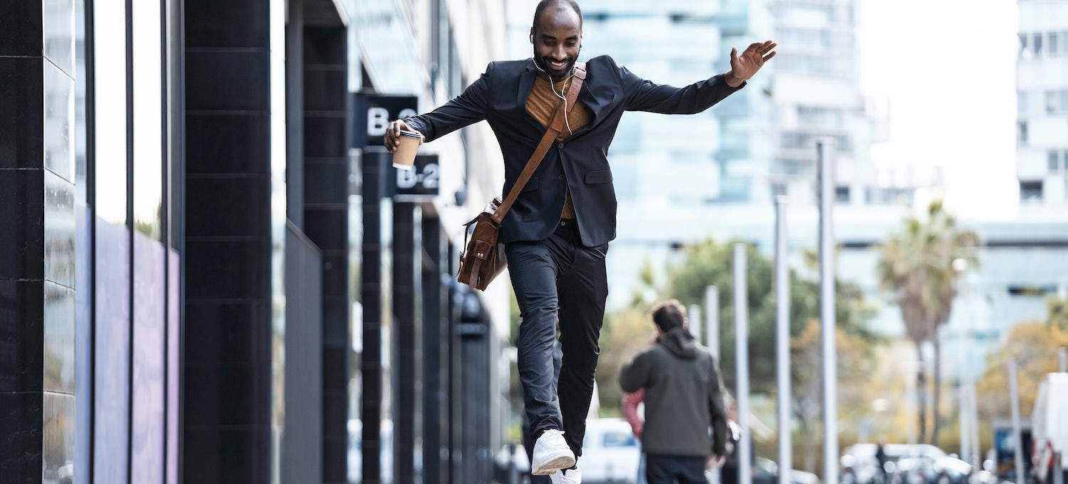 [Imagen destacada] Un hombre sonriente con un café salta de un banco a otro fuera de un edificio de oficinas.