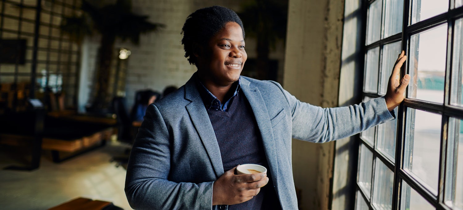 [Featured Image] A smiling person wearing a blazer and holding a coffee mug looks out the window.