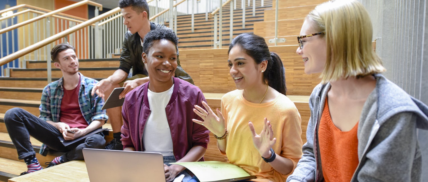 Three business school students sit on wooden steps in the foreground, with two more students sitting in the background, chatting and smiling in two separate groups as they refer to laptops and books in front of them.