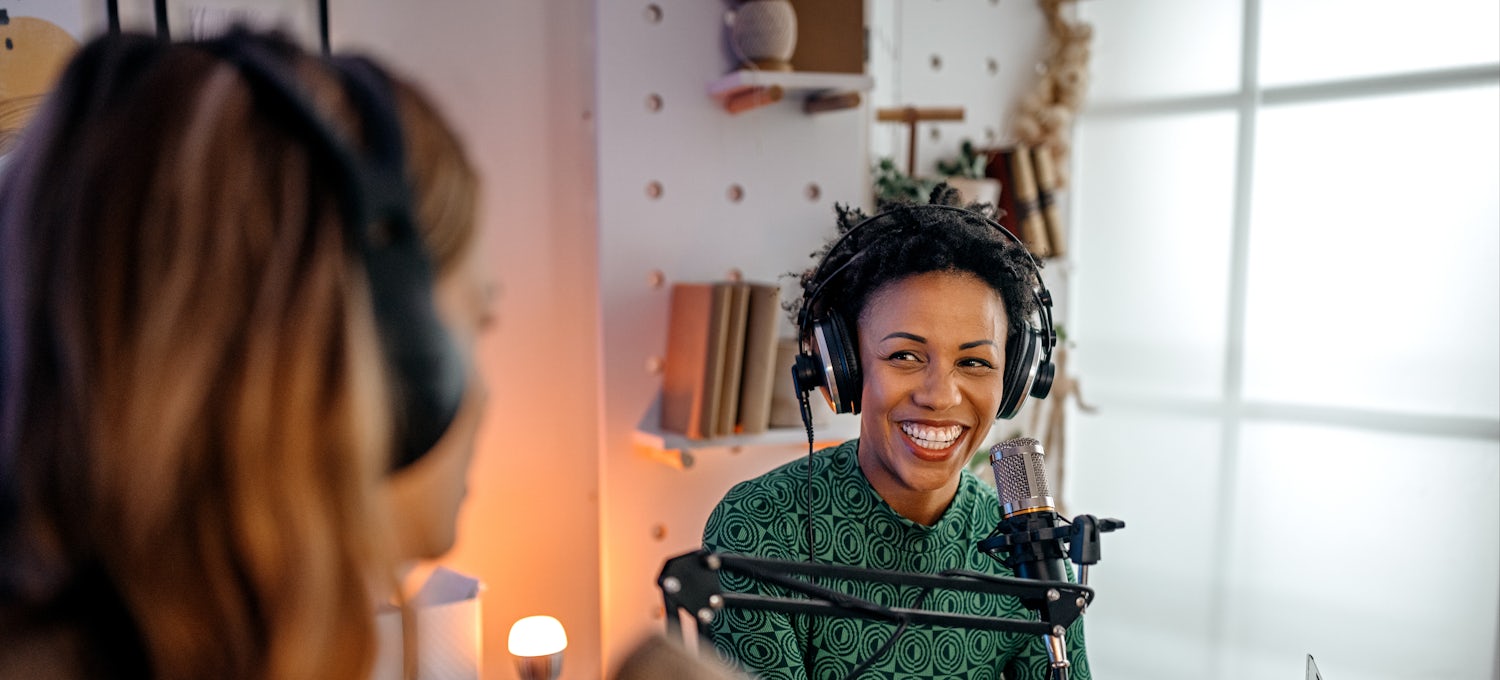 [Featured image] Two females, one wearing a green sweater and the second with long brown hair, are sitting in a room with a microphone wearing headphones on a desk, as they prepare to broadcast their cybersecurity podcast.