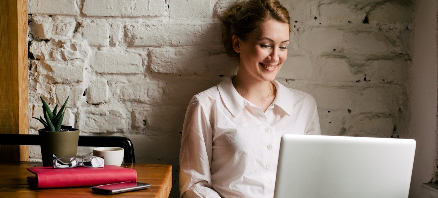 [Featured image] A virtual assistant wears a white blouse and works remotely on their laptop with a red planner and coffee cup to their right.