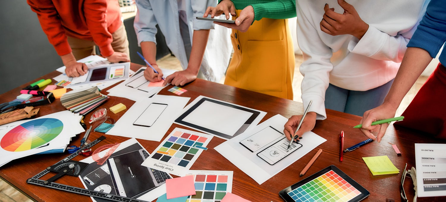 [Featured image] A group of digital designers reviews printouts of color schemes on a large table for mobile and tablet designs.