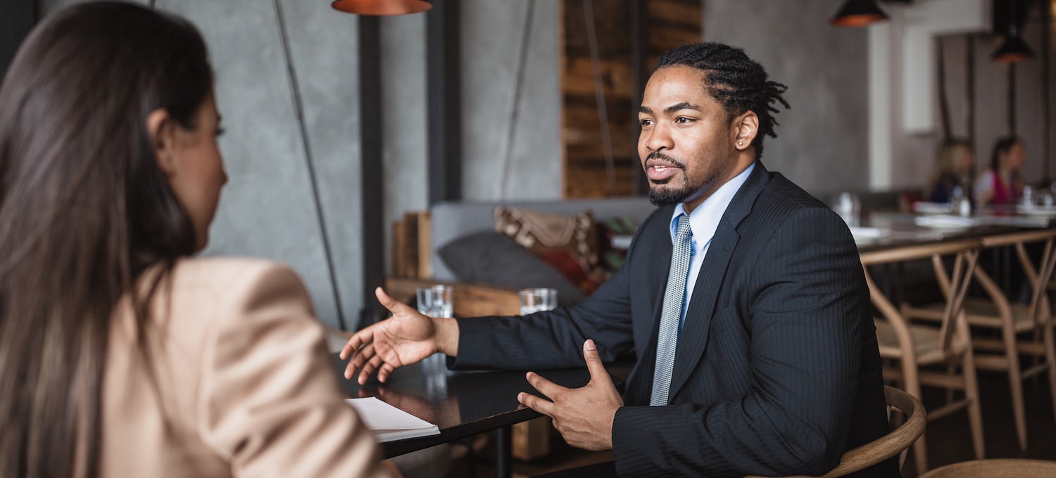 [Featured image] A young Black man in a business suit holds a conversation with a white woman in a business suit. 