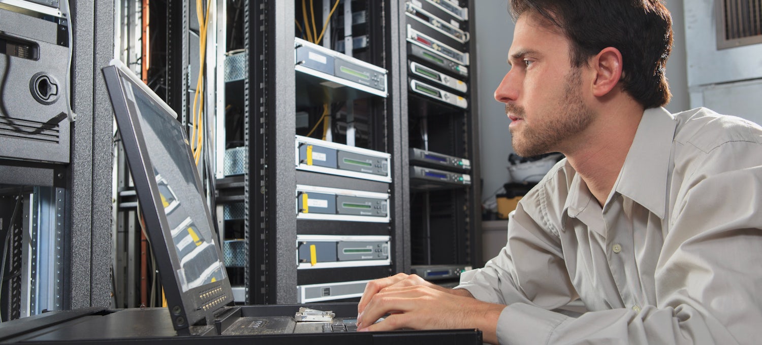 [Featured Image] A male network security engineer looking at a monitor in a cable server room and programming configurations.