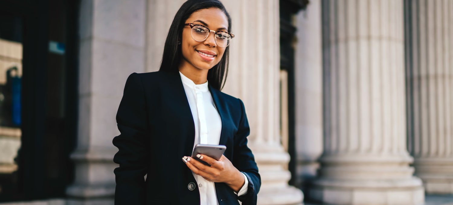 [Featured image] An MBA student stands outside a columned building holding her mobile phone and looking at the camera.