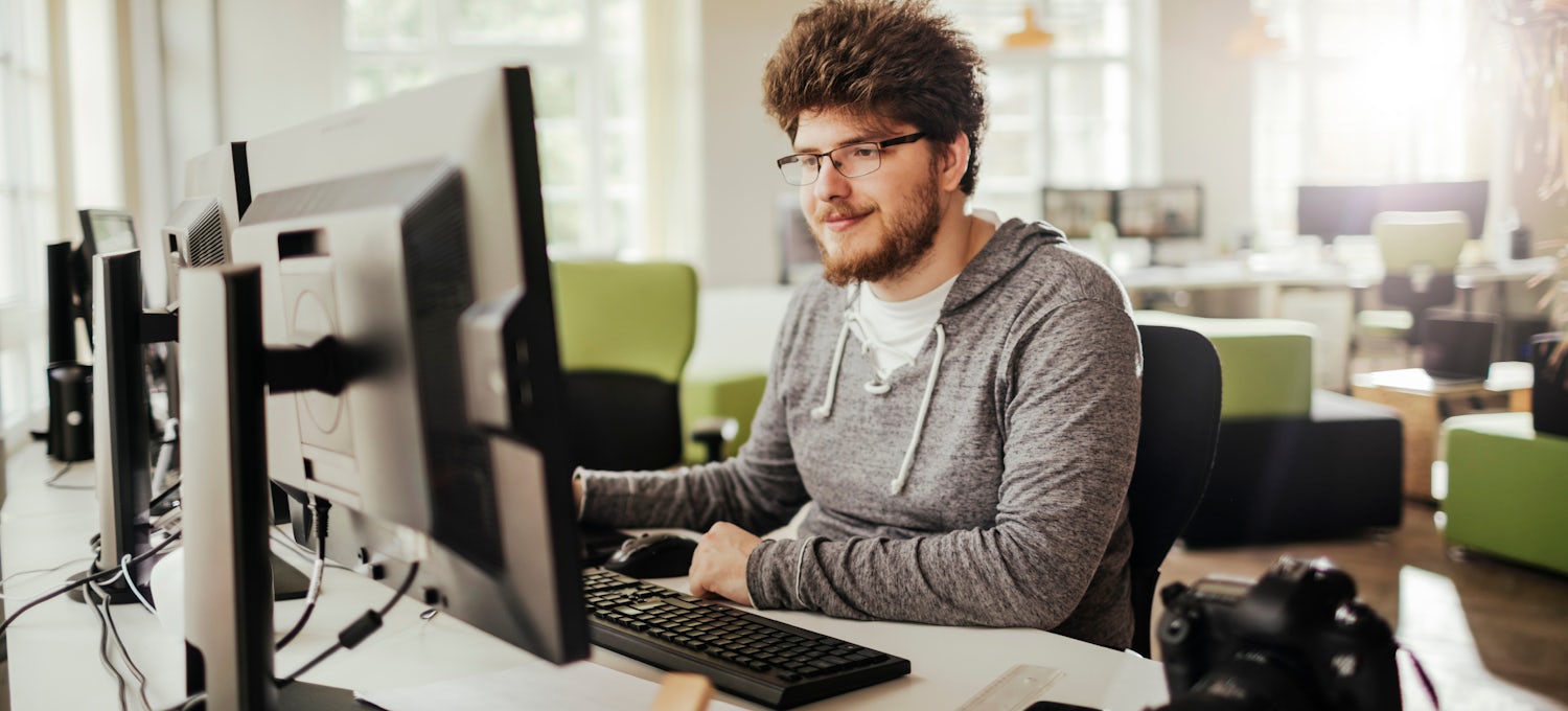 [Featured image] An AWS DevOps engineer in a gray hoodie and glasses sits at a desktop computer in a shared workspace.