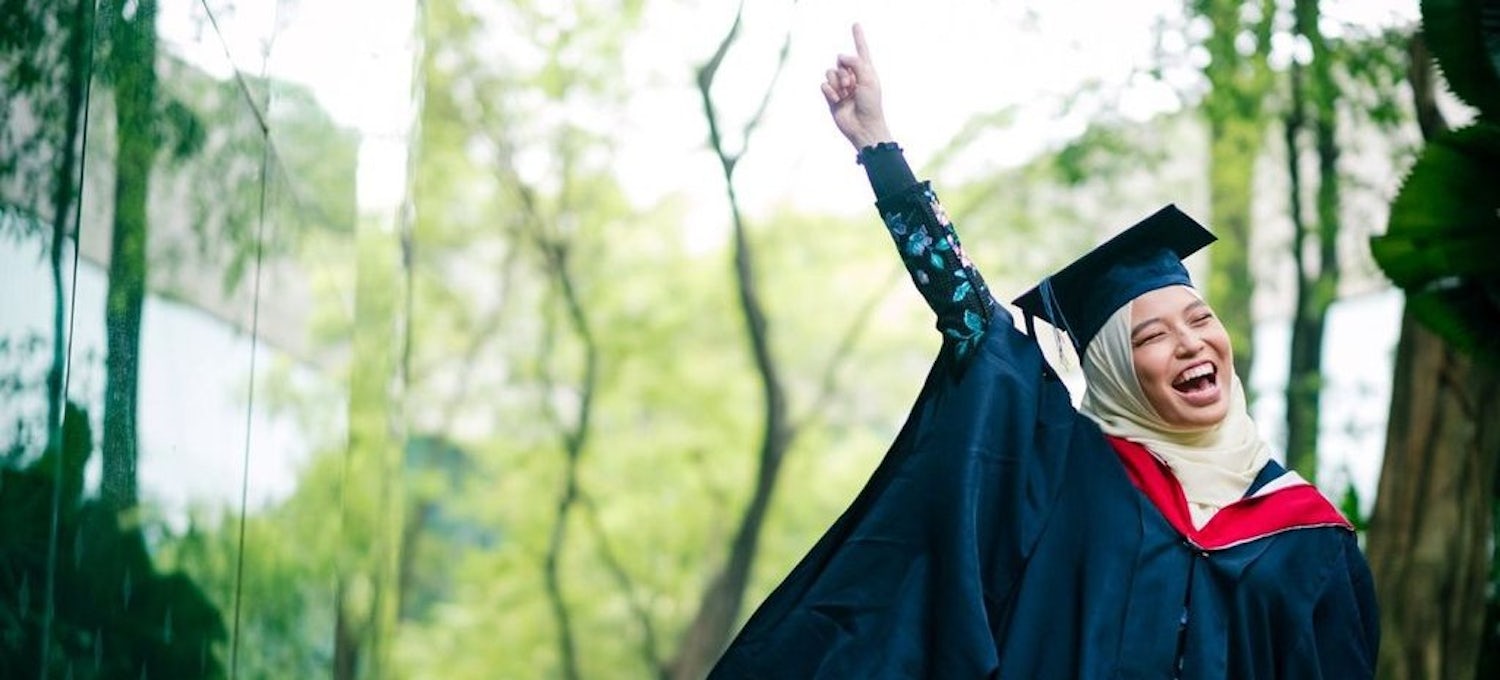 [Featured Image] A master's graduate student is in her black gown smiling and pointing to the sky.  