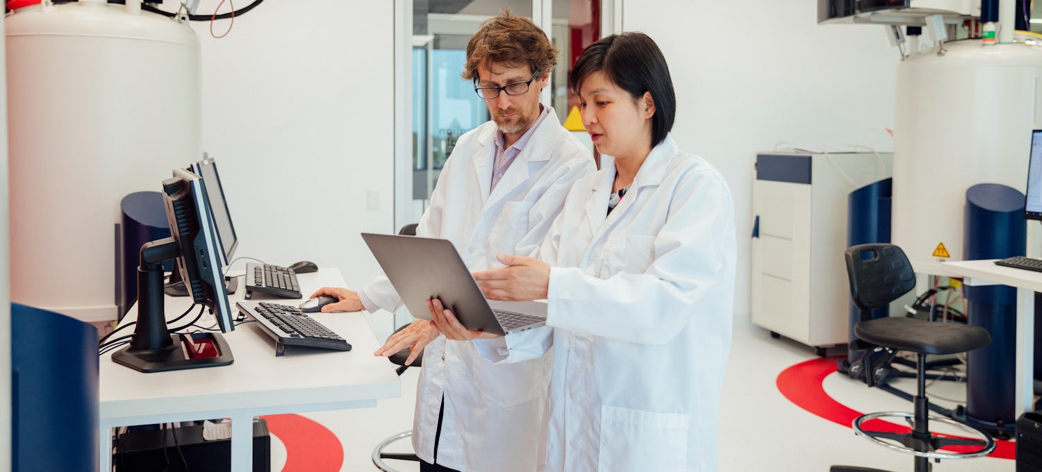 [Featured image] Two pharmacists in lab coats talk to each other while looking at a computer monitor.
