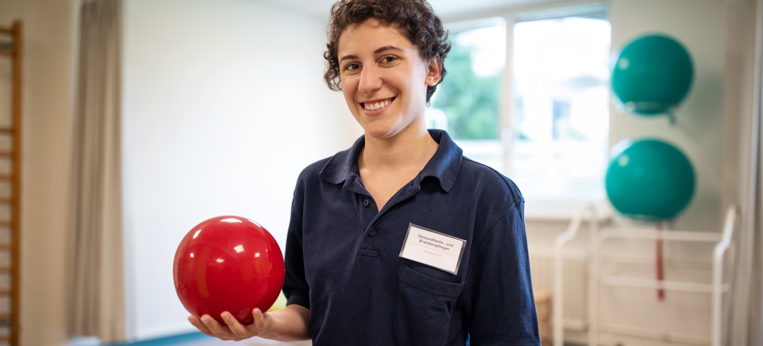 [Featured image] A physical therapist holding a red therapy ball stands in the middle of a clinic smiling at the camera.