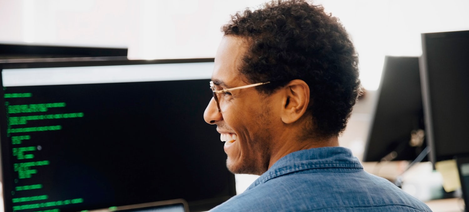 [Featured Image] A software engineer smiles in front of a computer monitor with code on the screen. 