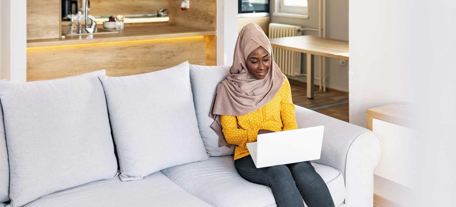 [Featured image] A woman wearing a hijab and a warm yellow sweater sits on her couch using her laptop.