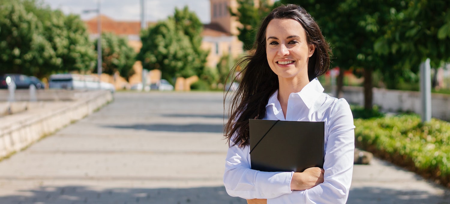 [Featured image] A young white woman with long brown hair, wearing a white collared shirt, stands on a college campus holding a folder. 
