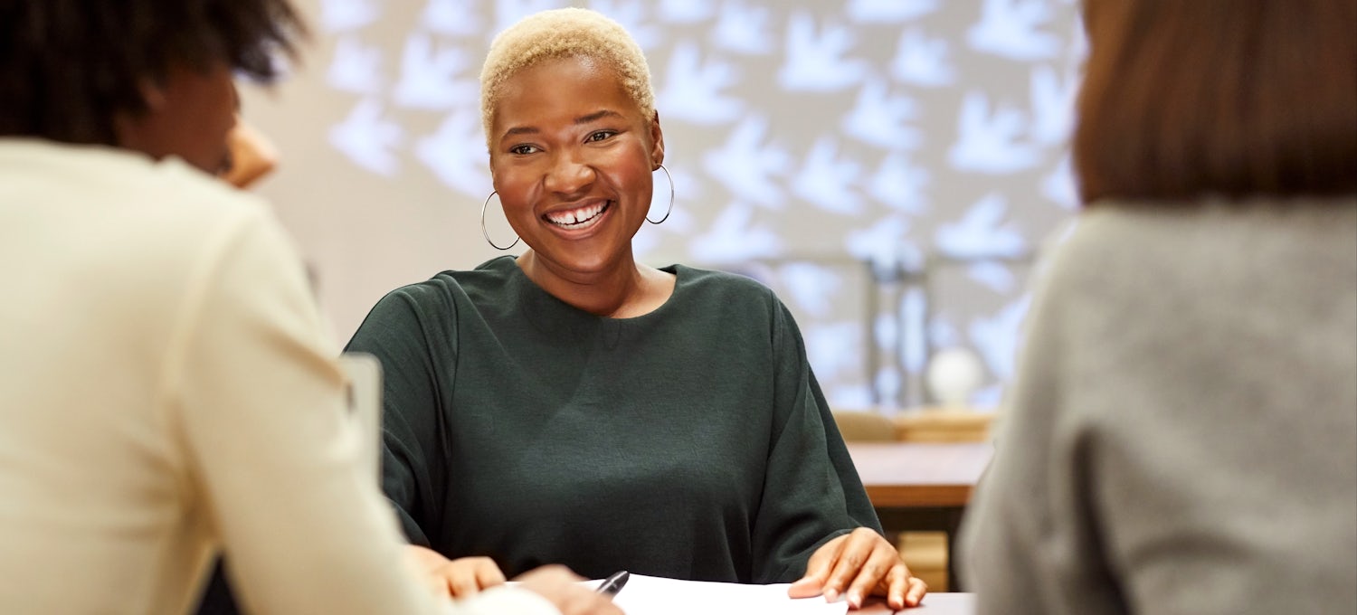 [Featured Image] Two black women interview another black woman who is smiling across the table with hoop earrings and green shirt.