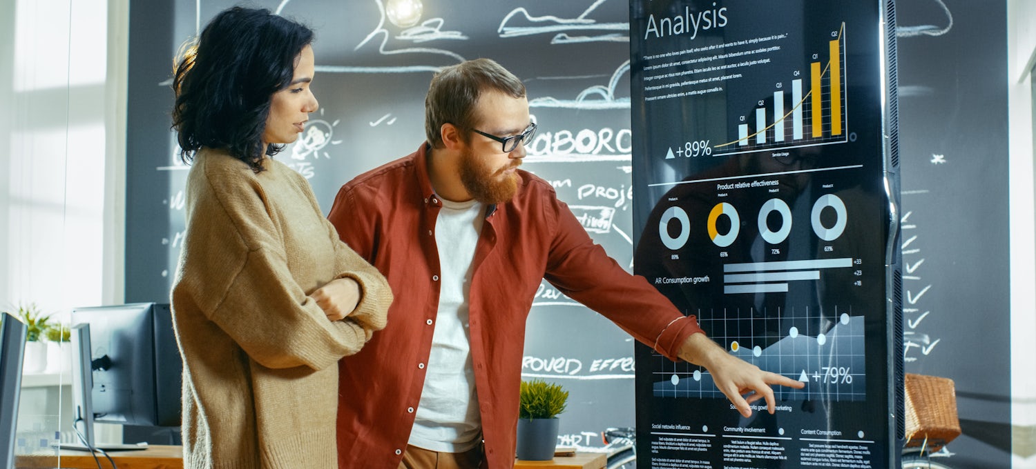 [Featured image] Two coworkers review sales forecasting data on a large screen display in an open workspace.