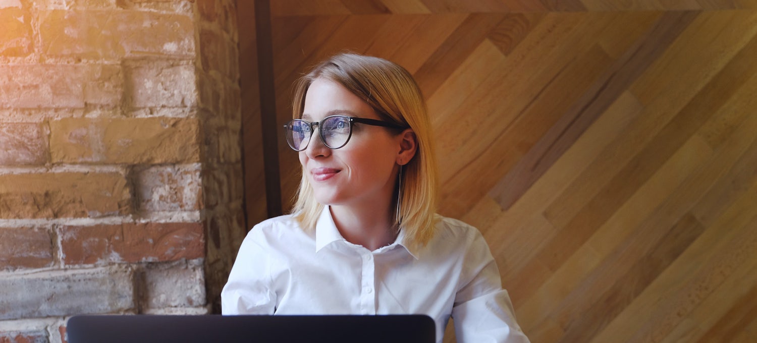[Featured image]:  A female,  with blonde hair, wearing a white shirt and glasses, is sitting in front of her desktop working on an assignment as she works towards her business administration degree.